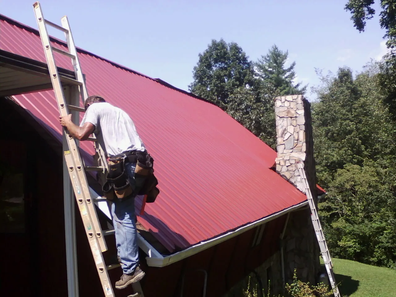 Roofer on a ladder beside a red metal roof and stone chimney — Soffit & Fascia Repair by Roofing Brothers in Rupert